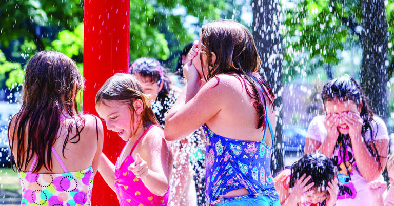 Several kids are playing in a splash pad, running and laughing under streams of water on a sunny day. They wear swimsuits and look happy as they cool off in the water.