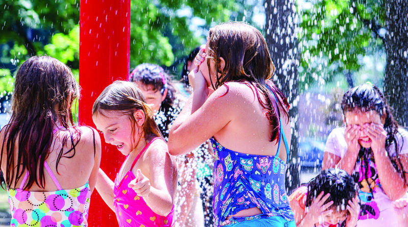 Several kids are playing in a splash pad, running and laughing under streams of water on a sunny day. They wear swimsuits and look happy as they cool off in the water.