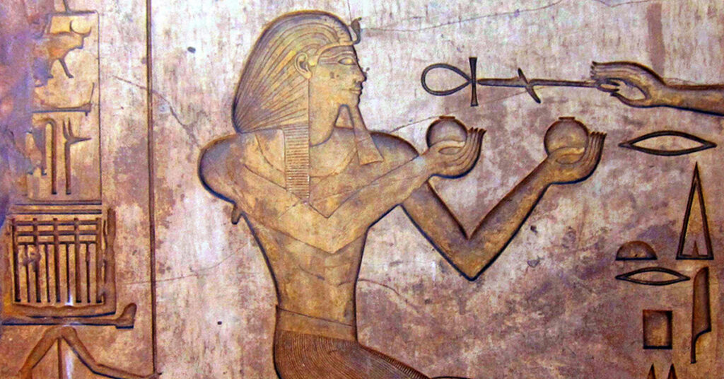 An ancient Egyptian carving shows a pharaoh kneeling, wearing a headdress, and holding two round objects in his hands. A mysterious hand extends an ankh, the symbol of life, toward him. Hieroglyphs and other symbols decorate the background.