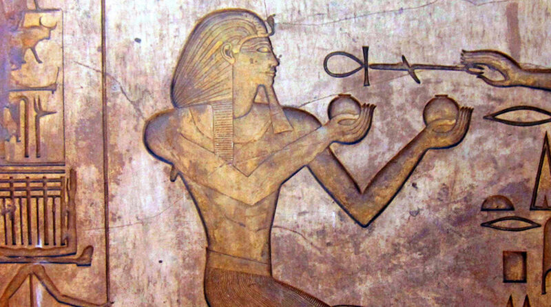 An ancient Egyptian carving shows a pharaoh kneeling, wearing a headdress, and holding two round objects in his hands. A mysterious hand extends an ankh, the symbol of life, toward him. Hieroglyphs and other symbols decorate the background.