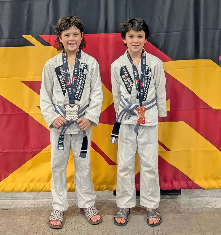 Two kids wearing jiu-jitsu uniforms smile and show off their medals in front of a red, black, and yellow championship banner.