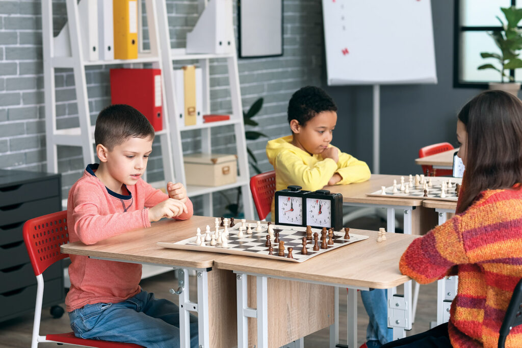 Four kids are playing chess on two tables in a classroom. There's a chess clock on each table.
