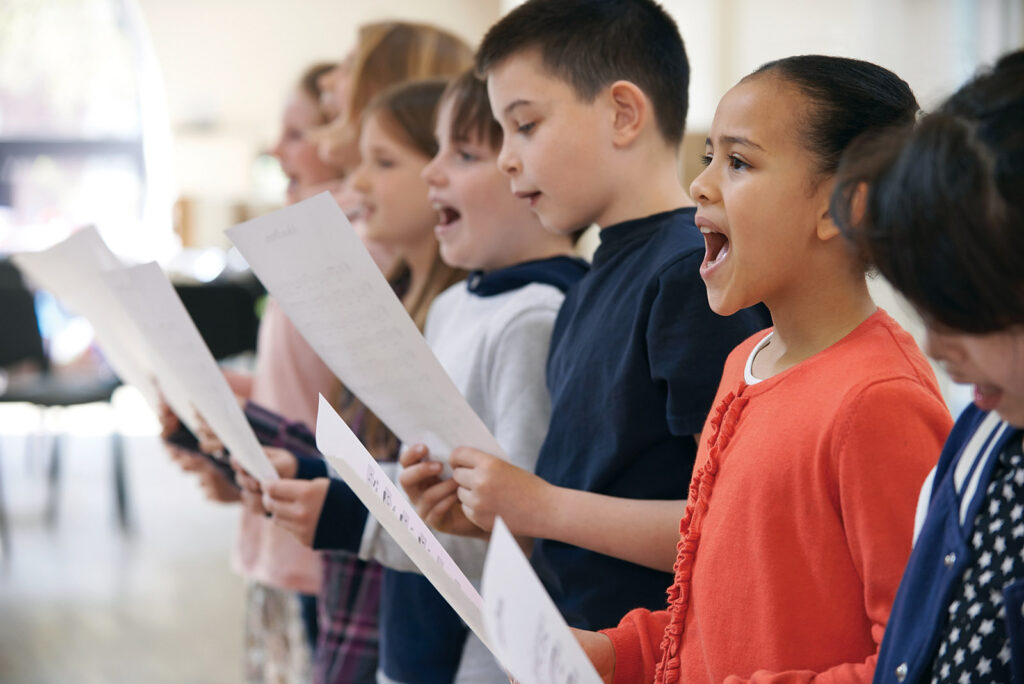 A group of kids singing together in a choir, holding sheet music and opening their mouths wide to sing. A girl in an orange shirt is in front.