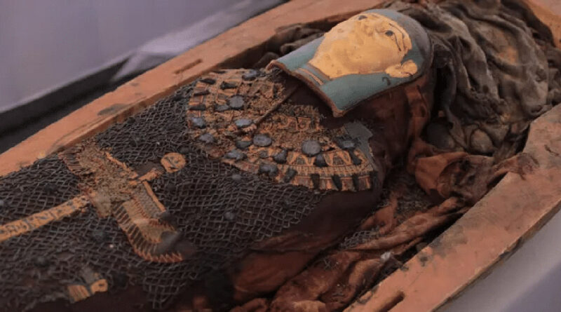 An ancient Egyptian mummy lies in a wooden coffin. The body is wrapped in cloth and decorated with beads, jewelry, and a painted mask of a face.