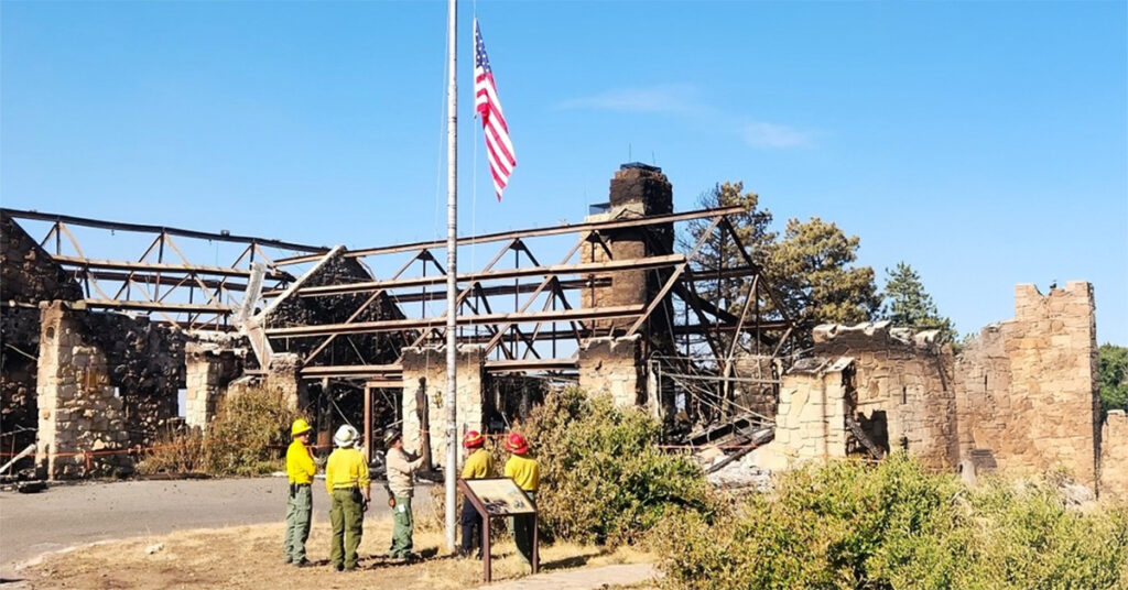 Firefighters stand near a flagpole with the U.S. flag at half-mast. Behind them is a burned building with only stone walls and roof beams left. The sky is clear and blue.