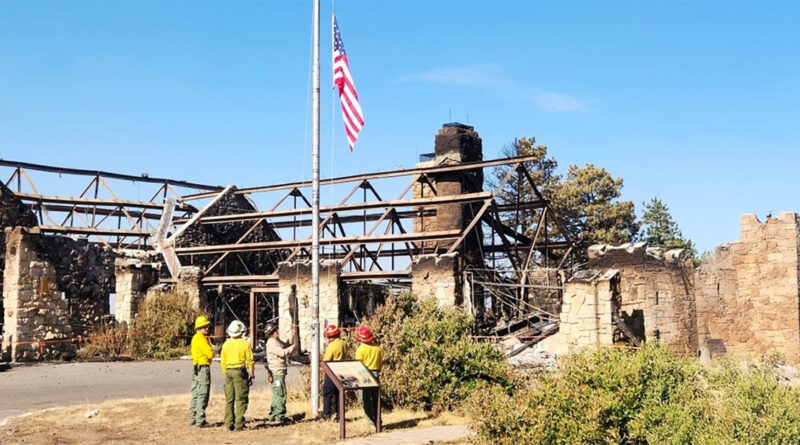 Firefighters stand near a flagpole with the U.S. flag at half-mast. Behind them is a burned building with only stone walls and roof beams left. The sky is clear and blue.