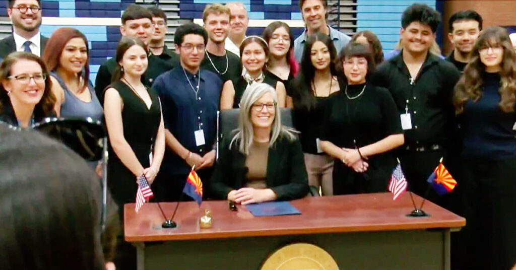A woman is sitting at a desk smiling with a group of students standing behind her. Small U.S. and Arizona flags are on the desk in front of her.