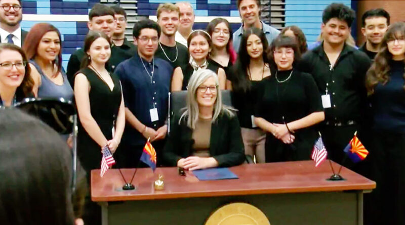 A woman is sitting at a desk smiling with a group of students standing behind her. Small U.S. and Arizona flags are on the desk in front of her.