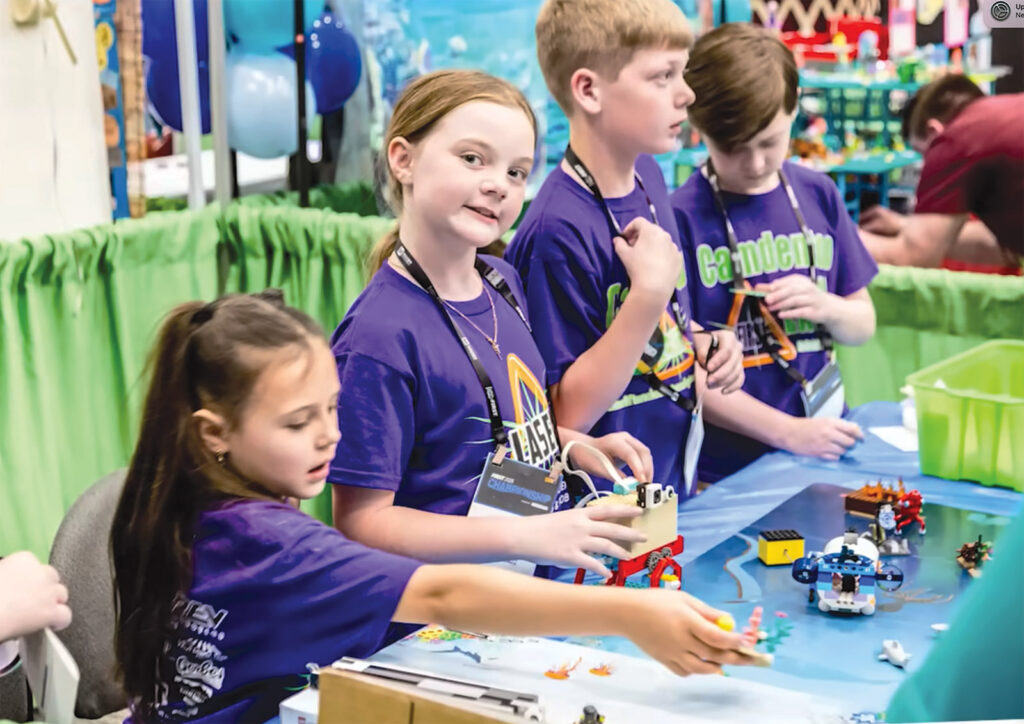 Kids in purple shirts building with LEGO blocks at tables during what looks like a robotics or STEM competition.