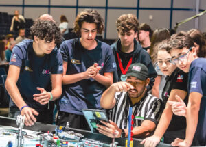 Teenage students in matching blue shirts gathered around a referee looking at a tablet during a robotics competition.