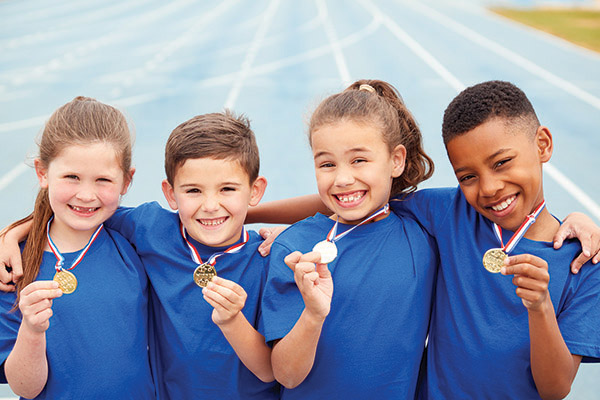 Four smiling kids in blue shirts are standing on a track. They each have medals around their necks and are holding them up proudly.