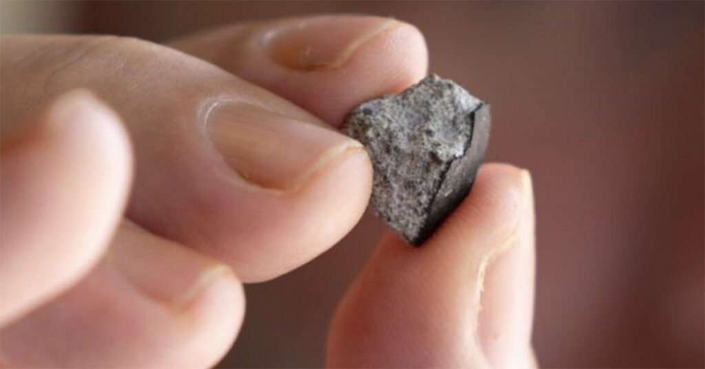 A small gray rock, about the size of a fingernail, is being held between two fingers. It’s rough and jagged and looks like it came from outer space.