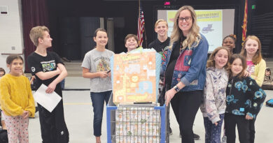A group of smiling kids stand with a woman holding a decorated box in a school gym.
