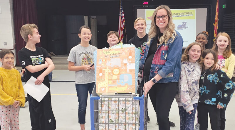 A group of smiling kids stand with a woman holding a decorated box in a school gym.