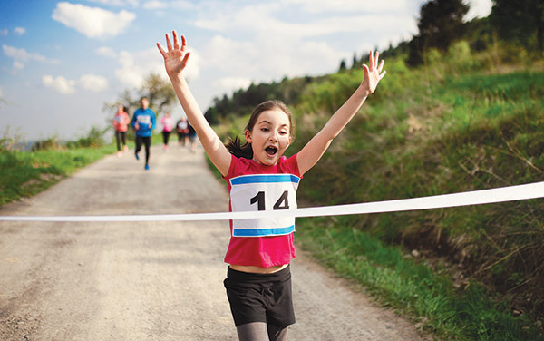 A young girl with race number 14 is running and smiling with her arms up as she crosses the finish line. Other runners are behind her on the dirt path.