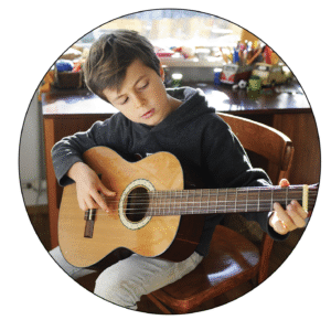 A young boy is sitting in a chair playing an acoustic guitar. He looks focused as he strums with one hand and presses the strings with the other.