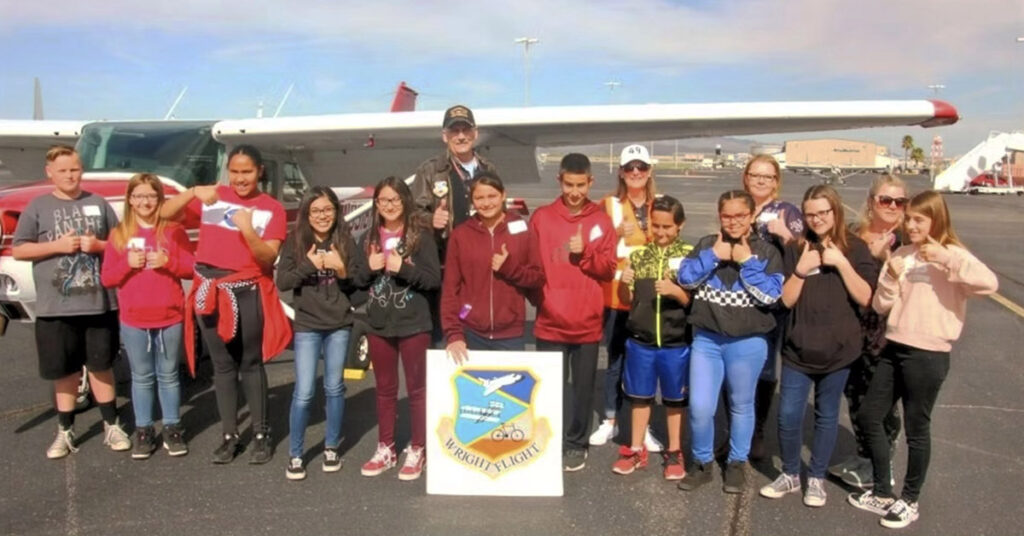 A large group of kids and teens posing with thumbs up in front of a small airplane on a runway with a Wright Flight sign.