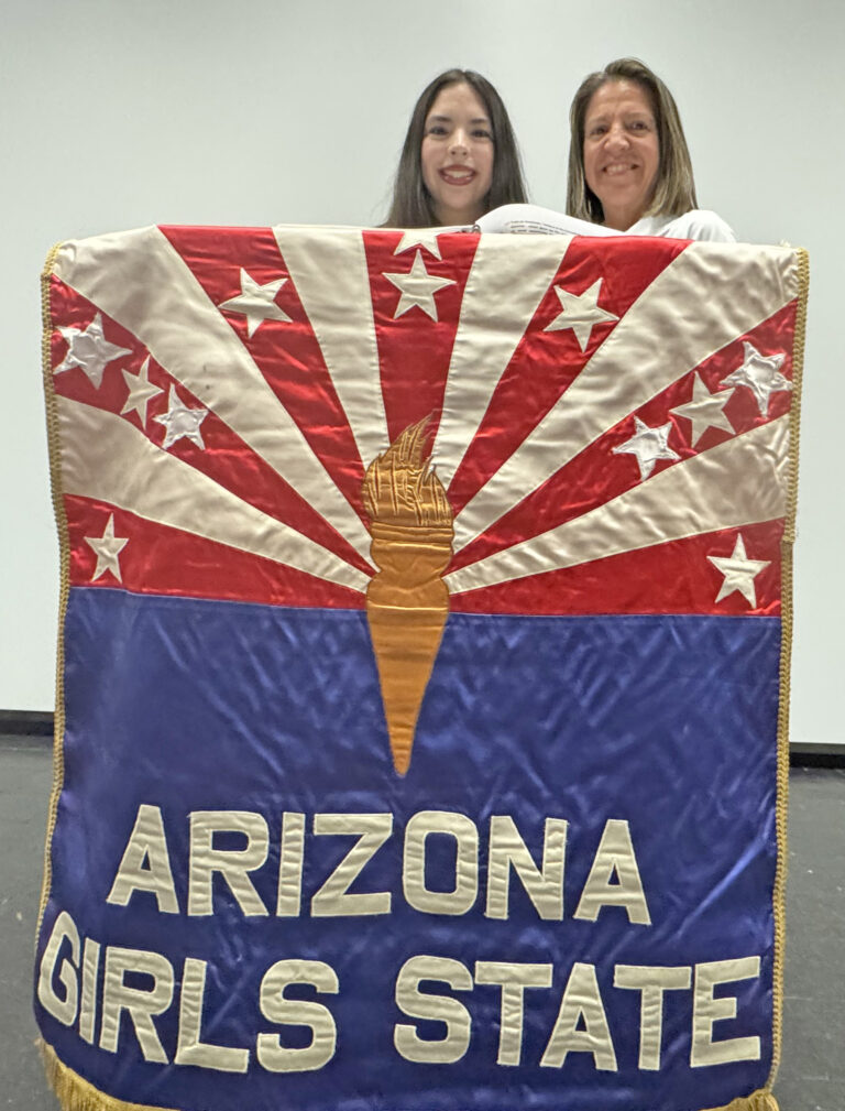 Two smiling women stand behind a large Arizona Girls State banner with red and white rays, white stars, a gold torch, and bold white letters on a blue background.