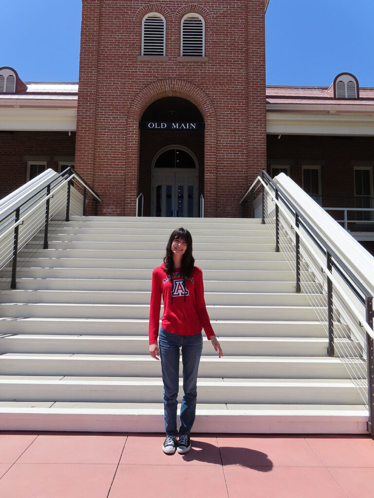 girl is standing and smiling in front of a big staircase at a red brick building that says “Old Main.” She’s wearing a red Arizona shirt and jeans.