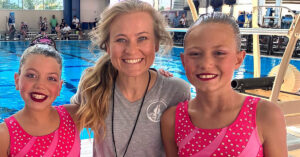 Two young girls in sparkly pink swimsuits and a smiling coach stand by an indoor pool, ready for a swimming event.