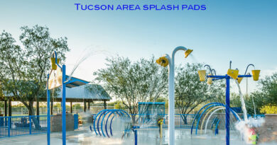 Colorful splash pad with water spraying from buckets, arches, and fountains on a sunny day, with trees and a shaded seating area in the background.