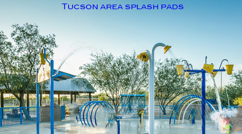 Colorful splash pad with water spraying from buckets, arches, and fountains on a sunny day, with trees and a shaded seating area in the background.