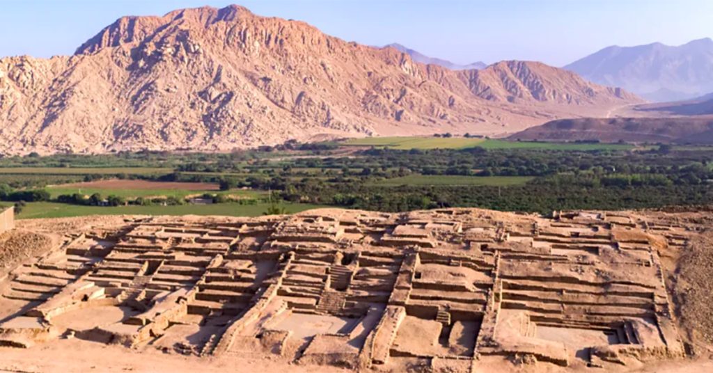 Stone ruins from an ancient civilization sit in a desert valley, surrounded by green farmland and tall mountains.