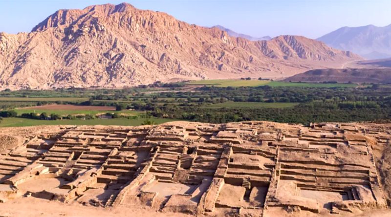 Stone ruins from an ancient civilization sit in a desert valley, surrounded by green farmland and tall mountains.