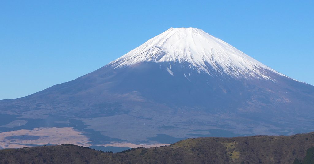 Snow-covered Mount Fuji stands tall under a bright blue sky, with green hills and fields in the foreground.