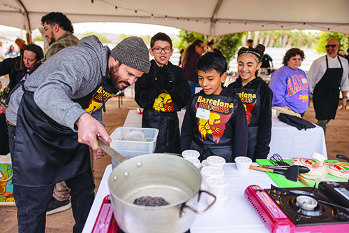 A chef shows three kids how to cook something in a pot at an outdoor cooking event under a big white tent.