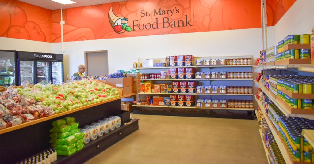 A food bank with shelves full of canned goods, boxed food, and fresh produce, with a person in a yellow vest helping inside.