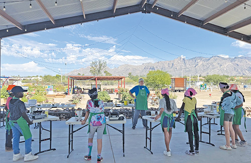 Kids wearing aprons stand at tables outside while a teacher teaches them about locally grown foods, with mountains in the background.