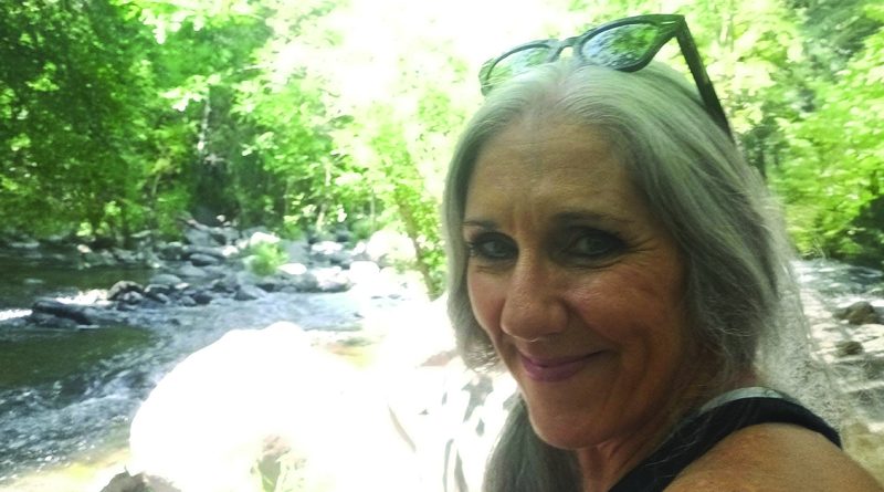 Smiling woman with gray hair and sunglasses on her head taking a selfie by a forest stream with green trees.