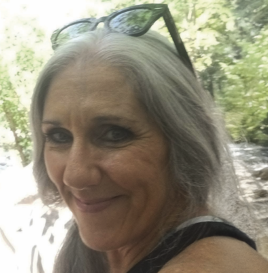 Smiling woman with gray hair and sunglasses on her head taking a selfie by a forest stream with green trees.