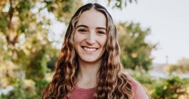 A woman with long curly hair smiles while standing outside. She wears a pink shirt. Trees are behind her.