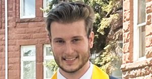 A young man in a dark blue graduation gown with a yellow sash smiles outside a brick building.