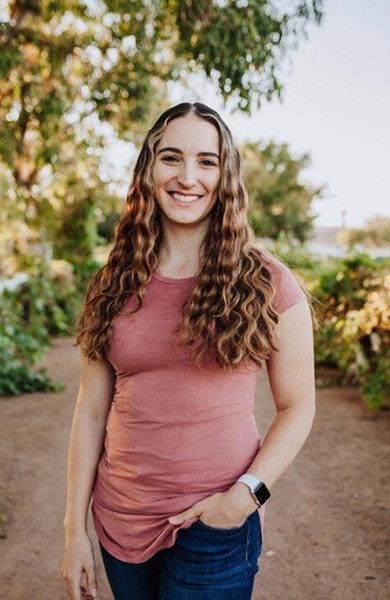 A woman with long curly hair smiles while standing outside on a dirt path. She wears a pink shirt, blue jeans, and a smartwatch. Trees are behind her.