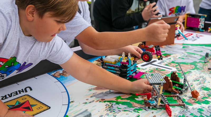 A boy leans over a table building a colorful LEGO machine on a big map while other kids work nearby.