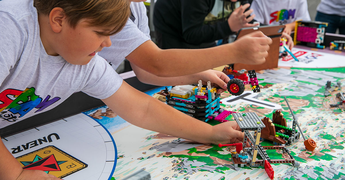 A boy leans over a table building a colorful LEGO machine on a big map while other kids work nearby.