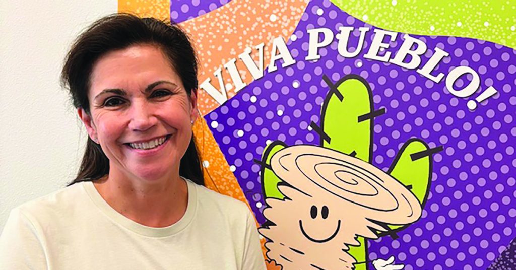 A smiling woman stands next to a colorful sign that says “Viva Pueblo!” with a happy cartoon cactus wearing a sombrero.