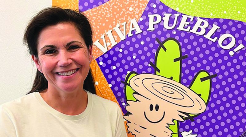 A smiling woman stands next to a colorful sign that says “Viva Pueblo!” with a happy cartoon cactus wearing a sombrero.