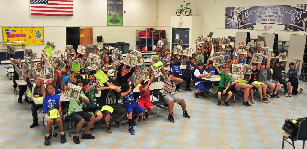Students in a school cafeteria cheer and hold up newspapers while sitting at tables during a reading activity.