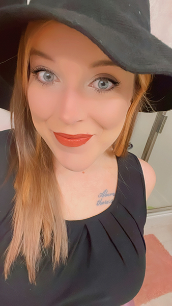 A woman with long light-brown hair smiles at the camera, wearing a black hat and black top, with bright eyes and orange-red lipstick.