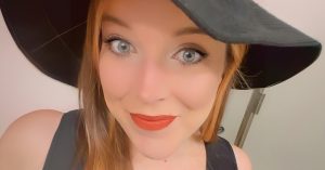 A woman with long light-brown hair smiles at the camera, wearing a black hat and black top, with bright eyes and orange-red lipstick.