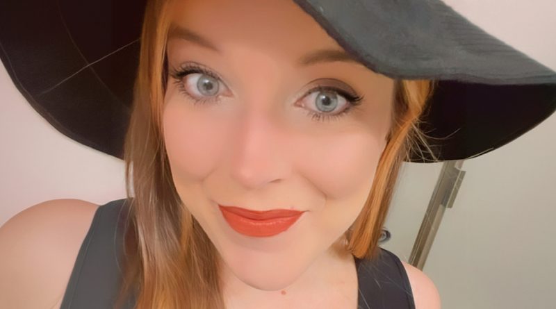 A woman with long light-brown hair smiles at the camera, wearing a black hat and black top, with bright eyes and orange-red lipstick.