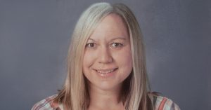 Smiling woman with long blonde hair wearing a striped shirt, photographed against a gray background in a professional headshot.