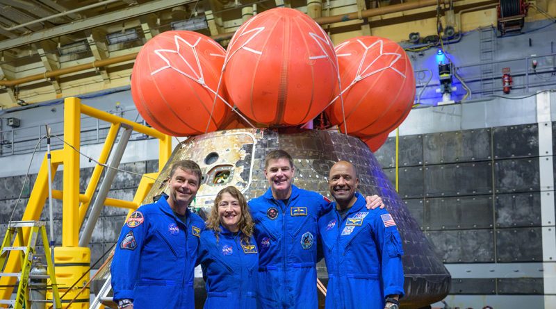 Four astronauts in blue NASA jumpsuits smile in front of a space capsule with orange parachutes in a large facility."