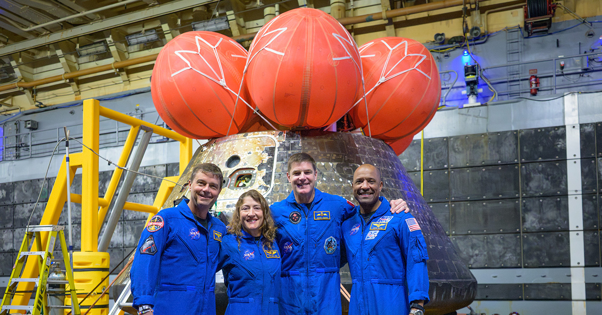 Four astronauts in blue NASA jumpsuits smile in front of a space capsule with orange parachutes in a large facility."