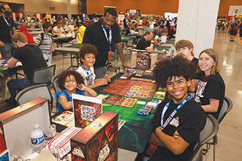 Group of kids and adults sitting at tables playing board games in a large convention hall with many people in the background.