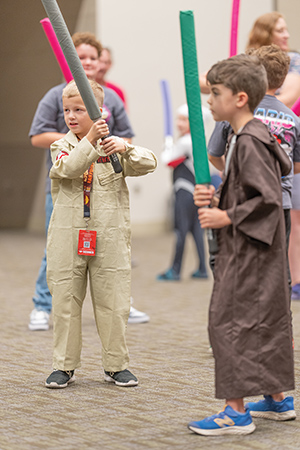 Kids dressed in costumes holding foam lightsabers at a convention, with one child in tan robes in the front.